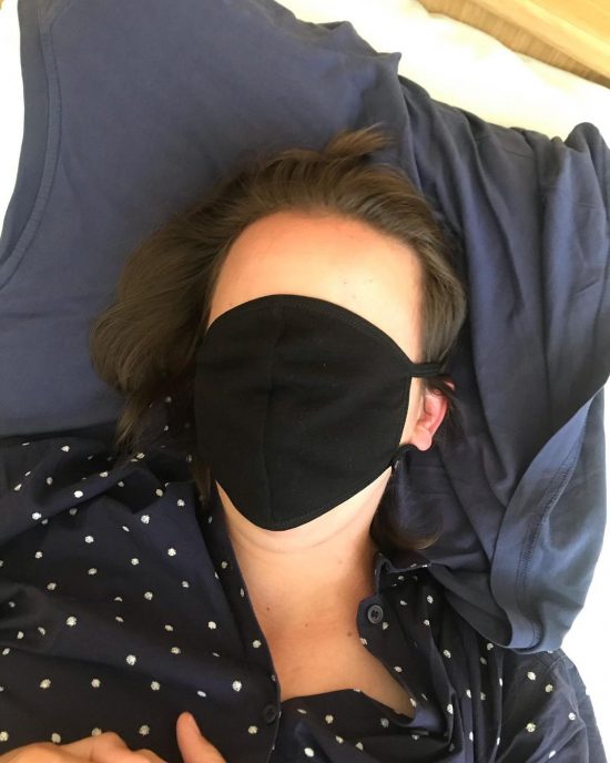 a figure laying on a navy blue pillow, over their face is a black face mask that, instead of covering the nose and mouth, also coviers their face, leaving thier forehead exposed. Their dark hair and dotted shirt lay crumpled on the pillow