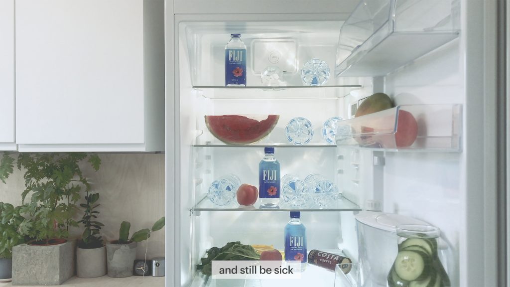 An image of a fridge door open, the contents curated - fija water bottle, water melon, apples, sliced cucumber in a glass water jug, the light from the fridge looks clean nad clinical. The caption below reads "this film didn't know it was sick"